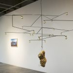 A mobile sculpture made of horizontal wires with bells on either end, and a center wire that holds a rock at the bottom, is in the foreground of the picture. Behind it is a gallery wall with a landscape photo.