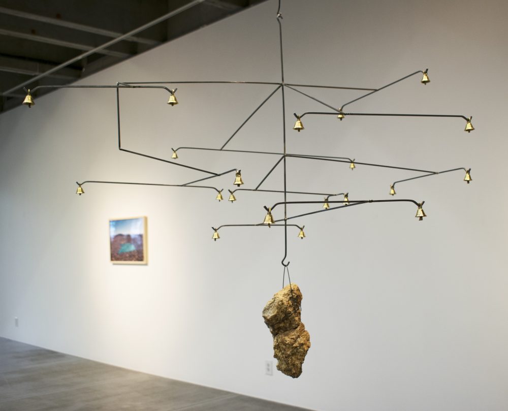 In the foreground is a mobile made of steel wires with brass bells, and a central wire with a rock hanging from it; a landscape photograph hangs on a wall in the background.