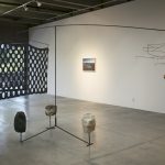 Large sculpture made with steel rods; 3 large rocks are at the base; it has a wide screen with large holes on the left, and a wire mobile with bells on the right. In the background is a wall with a landscape photo hanging on it.