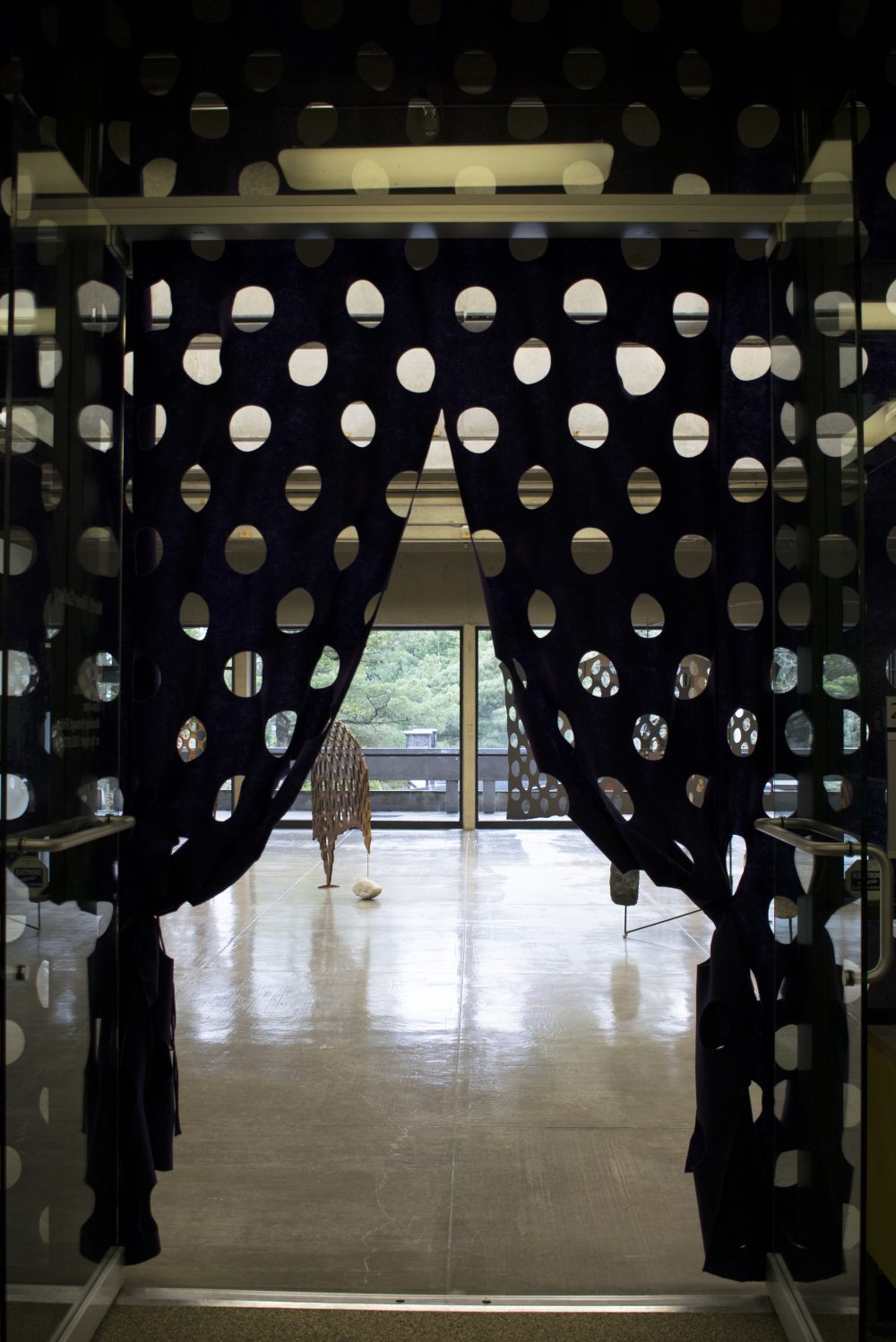 A dark curtain with a grid of large holes hangs in the foreground, it is parted in the middle and tied off on either side and reveals a gallery with sculptures inside.