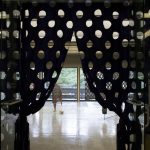 A dark curtain with a grid of large holes hangs in the foreground, it is parted in the middle and tied off on either side and reveals a gallery with sculptures inside.
