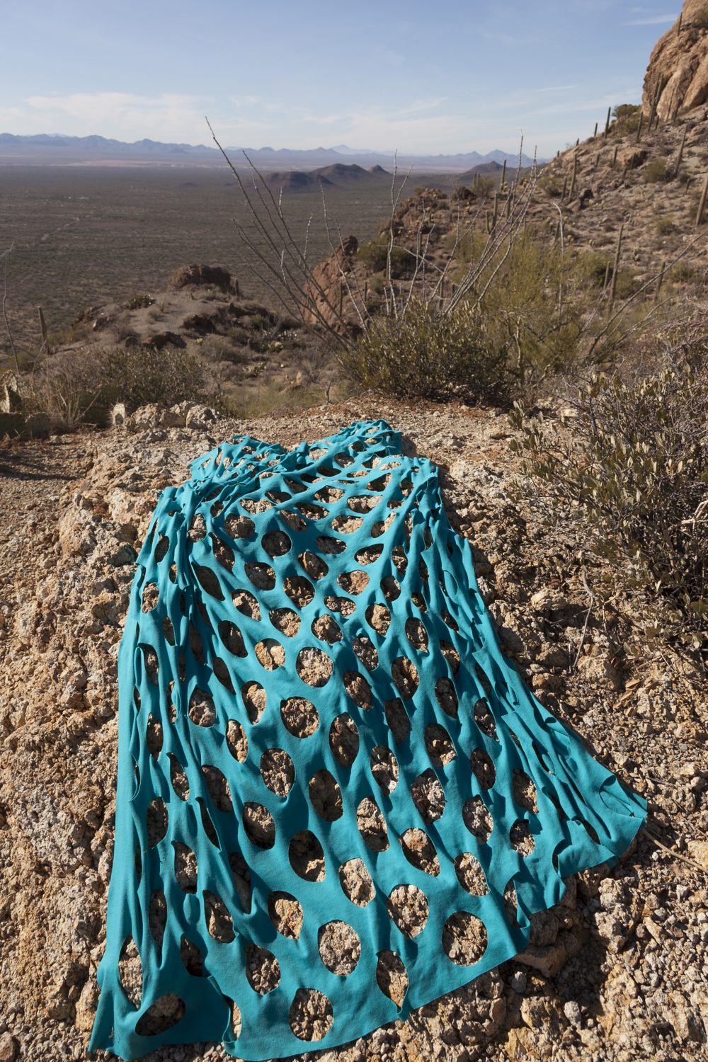 Color photograph of a desert landscape showing distant hills and sky in the background on the upper left of the image. In the foreground is a rectangular blue fabric with a grid of large holes cut out of it.