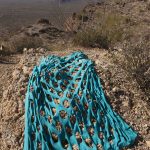 Color photograph of a desert landscape showing distant hills and sky in the background on the upper left of the image. In the foreground is a rectangular blue fabric with a grid of large holes cut out of it.
