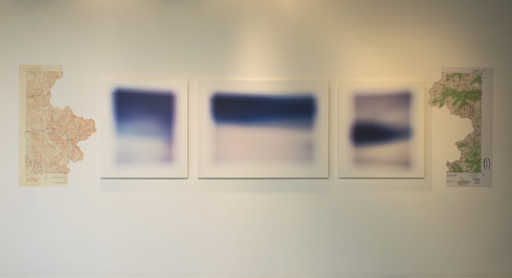 3 dark blue abstract landscape photos hung on a gallery wall, with 2 shaped map photos on either side.