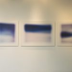 3 dark blue abstract landscape photos hung on a gallery wall, with 2 shaped map photos on either side.