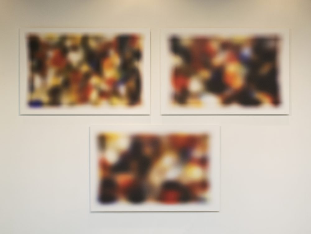 3 abstract, multicolored rectangular photos, hung on a gallery wall, 2 on top and 1 below.