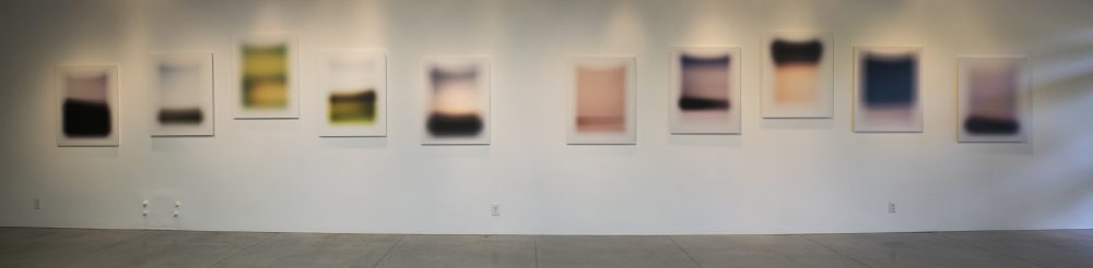 Wall of gallery with 10 photographs of simple abstract landscapes that are arranged in a wave pattern.