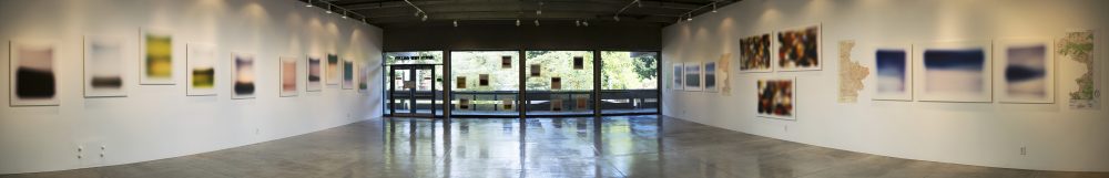 Panorama view of gallery with abstract photos on left and right sides, and in the middle the floor to ceiling windows with photos mounted on the glass and a view of trees outside.