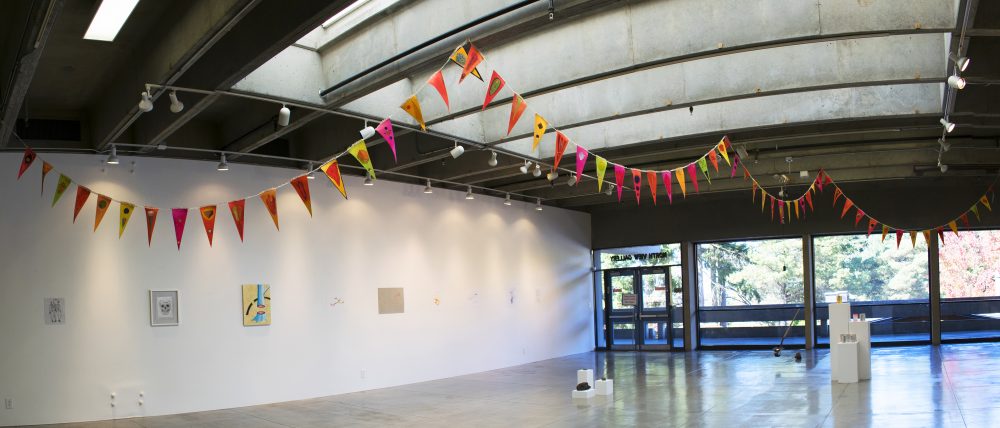 Gallery installation view of artwork made up of triangular flags attached to a ribbon and draped across the ceiling of the gallery; the flags are yellow, orange and pink, with various painted images on them. The gallery has other artworks throughout and in the background are large windows showing the trees outside.