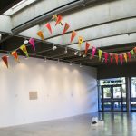 Gallery installation view of artwork made up of triangular flags attached to a ribbon and draped across the ceiling of the gallery; the flags are yellow, orange and pink, with various painted images on them. The gallery has other artworks throughout and in the background are large windows showing the trees outside.