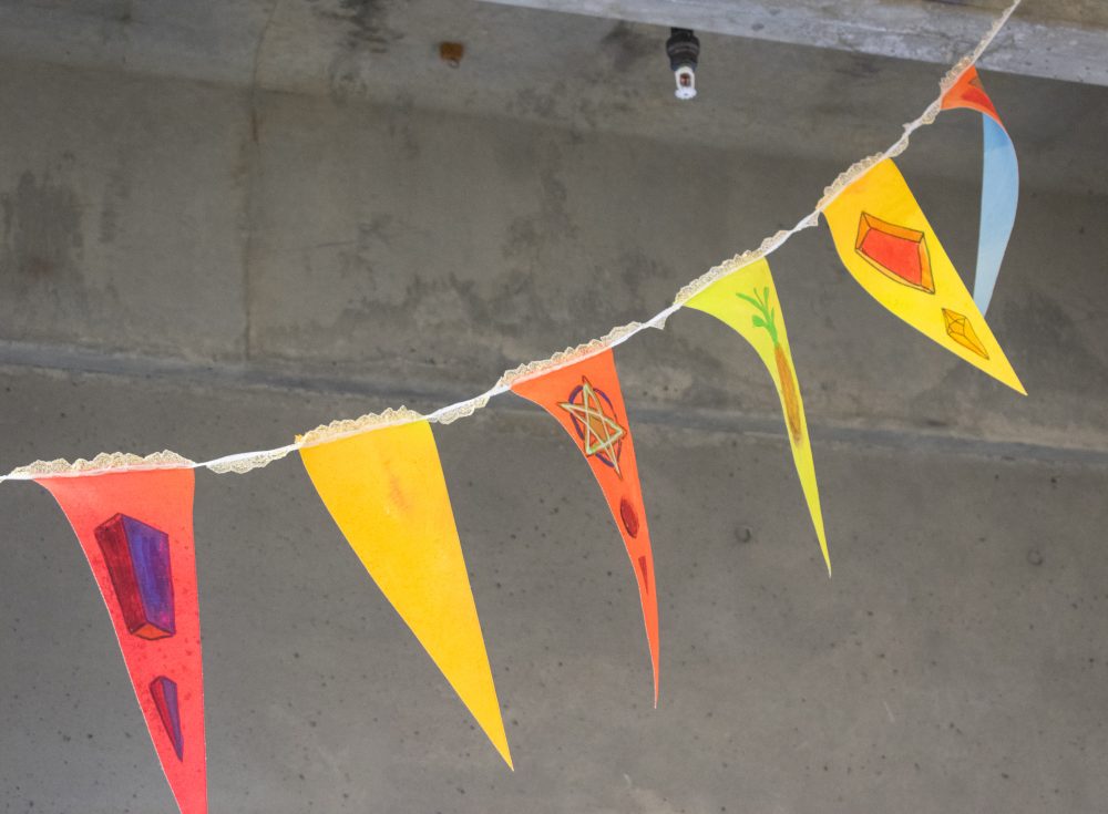 White ribbon with five triangular flags attached to it. The yellow, orange and pink flags have abstract images painted on them; the background is a concrete ceiling.