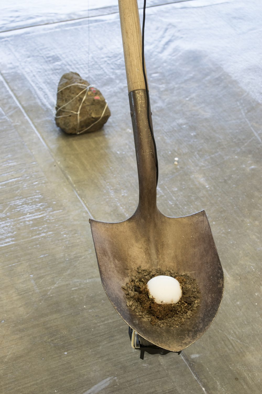 Close up view of shovel with ball of ice surrounded by moss sitting in blade, a large stone sits on floor behind the shovel.