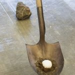 Close up view of shovel with ball of ice surrounded by moss sitting in blade, a large stone sits on floor behind the shovel.