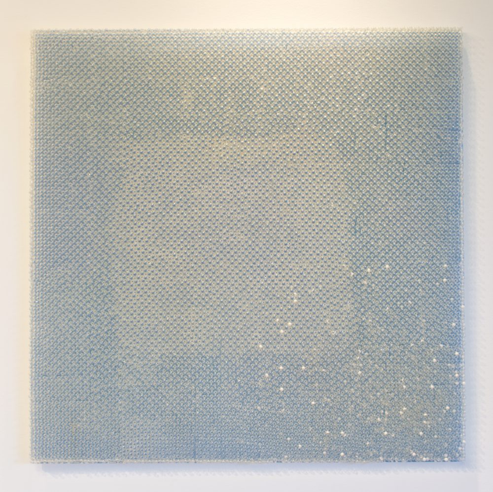Square, wall mounted artwork made of gel caps (pills); the design is a bluish outer square and a lighter whitish square inside; the surface is very reflective.
