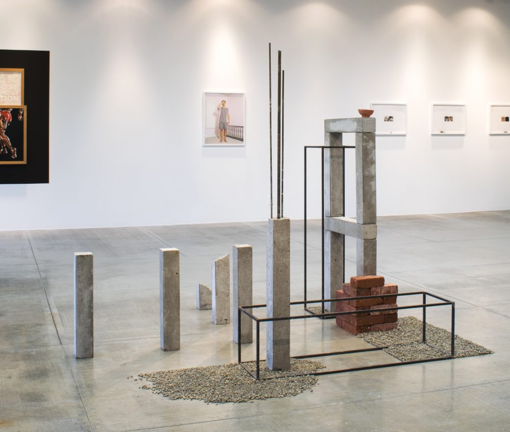 Abstract, geometric sculpture made up of an arrangement of concrete columns, gravel, bricks and clay; it sits on the floor of a gallery with other artworks on the wall in the background.