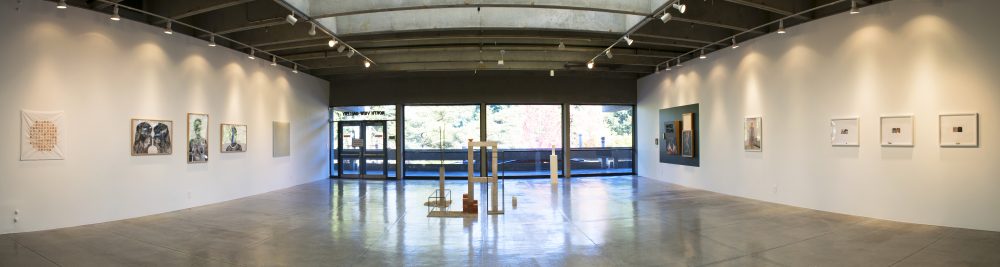 Panorama view of gallery with five artworks on left wall, five artworks on right wall and sculpture in the center, with large windows in the background showing the trees outside.