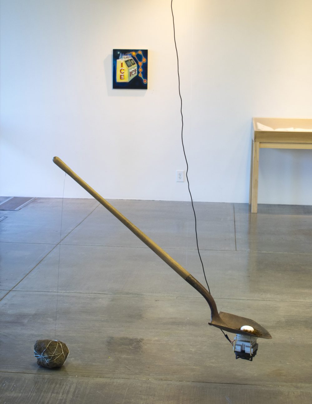 A shovel is suspended from the ceiling with a thermoelectric cooler attached to the underside of the blade and a ball of ice on the top side of the blade. A wire goes from the cooler to the ceiling, a large stone sits on the floor behind the shovel, a painting and a table are at the wall in the backgroundl.
