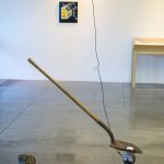 A shovel is suspended from the ceiling with a thermoelectric cooler attached to the underside of the blade and a ball of ice on the top side of the blade. A wire goes from the cooler to the ceiling, a large stone sits on the floor behind the shovel, a painting and a table are at the wall in the backgroundl.