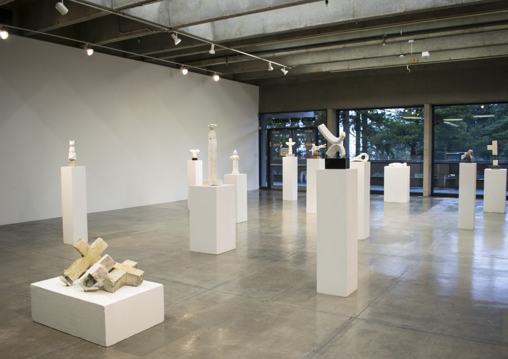 View of gallery with several marble sculptures sitting on pedestals. In the back of the gallery are large windows with views of trees.