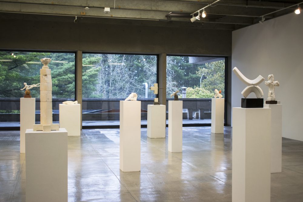 View of gallery with several marble sculptures on pedestals. In the background are large windows with views of trees.