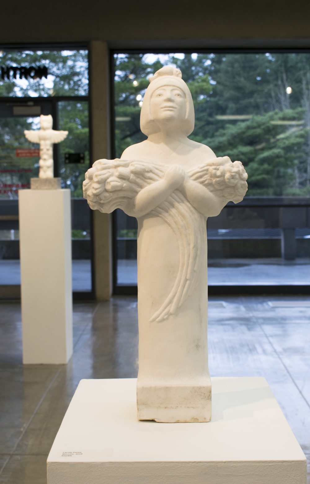 Marble sculpture of woman standing and holding flowers in arms that are across her chest. This sculpture is on a pedestal, in the background is another sculpture on a pedestal.