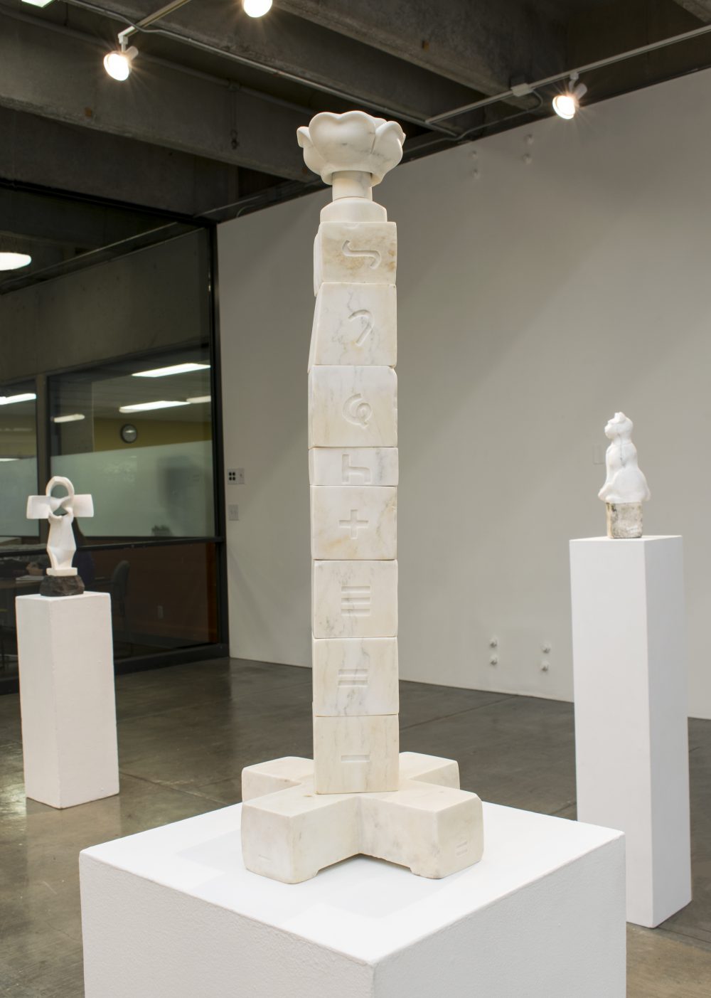 Marble sculpture consisting of cubes with incised letter forms stacked to form a 33 inch tower. On top is a flower form and on bottom, a cross form, Other sculptures are in the background.