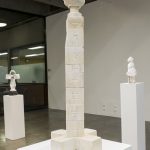 Marble sculpture consisting of cubes with incised letter forms stacked to form a 33 inch tower. On top is a flower form and on bottom, a cross form, Other sculptures are in the background.