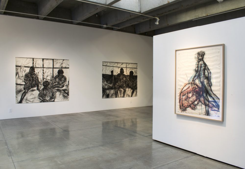 Two large charcoal drawings of three girls in silhouette by a window hang on wall on left of image, and one framed charcoal and wathercolor drawing of two female figures hangs on another wall on the right of the image.
