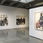 Two large charcoal drawings of three girls in silhouette by a window hang on wall on left of image, and one framed charcoal and wathercolor drawing of two female figures hangs on another wall on the right of the image.