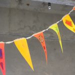 White ribbon with five triangular flags attached to it. The yello, orange and pink flags have abstract images painted on them; the background is a concrete ceiling.