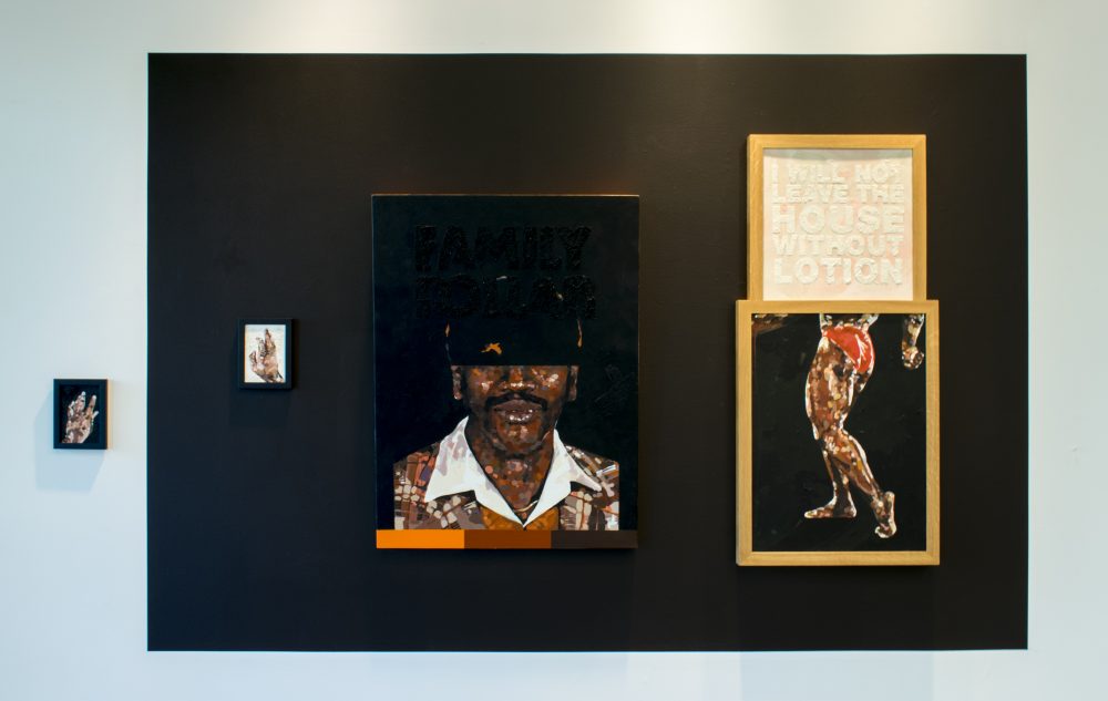 Black rectangle painted on wall; one small painting of a hand his on the left of the rectangle; 3 paintings hang within the rectangle: a small one of a hand, a larger painting of an African American man's face, half concealed, and another of a person's legs, with a framed text piece placed on top of that.