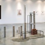 Abstract, geometric sculpture made up of an arrangement of concrete columns, gravel, bricks and clay; it sits on the floor of a gallery with other artworks on the wall in the background.