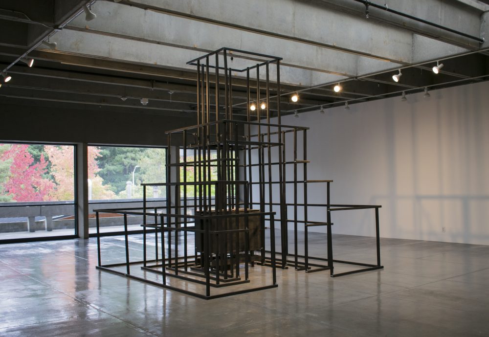 Large scale sculpture made of wood, in a pyramidal form, resembling a jungle jim play structure. Wood is painted black; in the center of framelike structure is a wooden box.