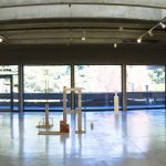 view of gallery installation with 5 artworks on left wall, 6 artworks on right wall, and 2 sculptures on floor in between. In the background are large windows with trees.
