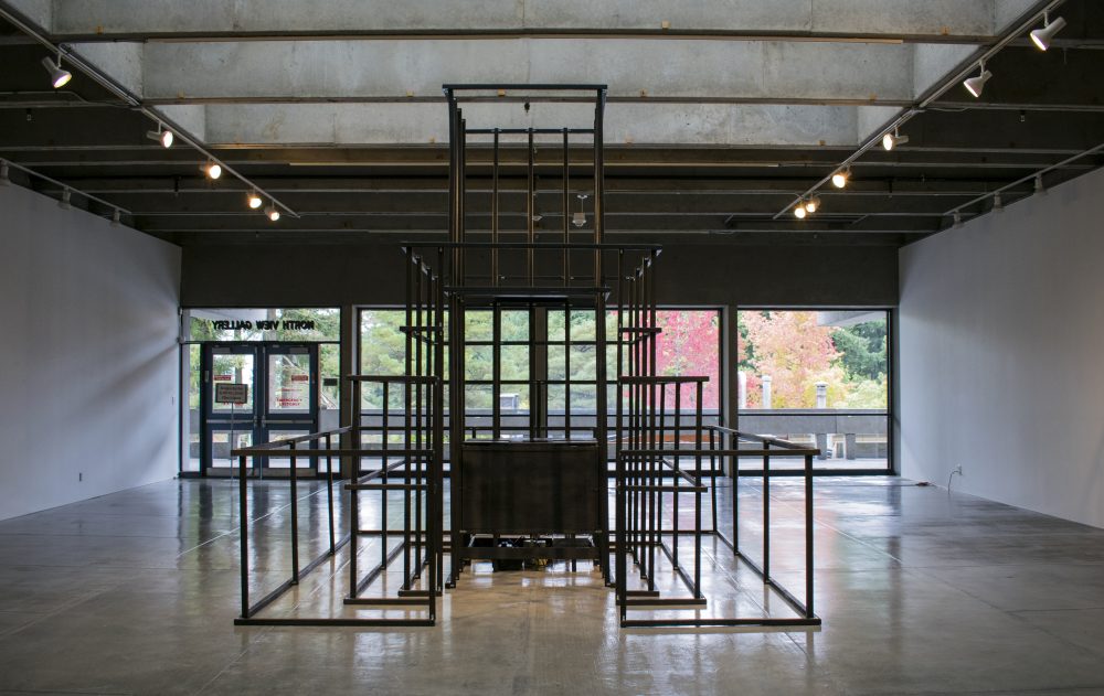 Large scale sculpture made of wood, in a pyramidal form, resembling a jungle jim play structure. Wood is painted black; in the center of framelike structure is a wooden box.