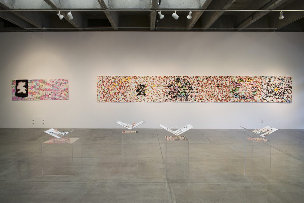 View of gallery with 2 long, horizontal, abstract paintings on a wall in the background and 4 plexiglass pedestals with folded paper artworks in the foreground. The painting on the left has several cut out circles and a larger cut out form; the one on the right has an overall complex patterning and is mostly red, black and white.