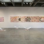 View of gallery with 2 long, horizontal, abstract paintings on a wall in the background and 4 plexiglass pedestals with folded paper artworks in the foreground. The painting on the left has several cut out circles and a larger cut out form; the one on the right has an overall complex patterning and is mostly red, black and white.