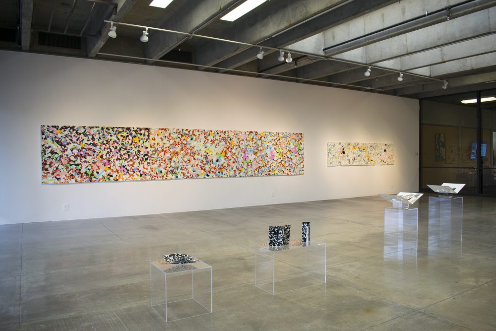 View of gallery with 2 long, horizontal, abstract paintings on wall in the background and 4 plexiglass pedestals with folded paper artworks in the foreground. Both paintings have an overall complex patterning; the one on the right is mostly black, red, and white; the one on the left is mostly white and grey.