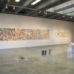 View of gallery with 2 long, horizontal, abstract paintings on wall in the background and 4 plexiglass pedestals with folded paper artworks in the foreground. Both paintings have an overall complex patterning; the one on the right is mostly black, red, and white; the one on the left is mostly white and grey.