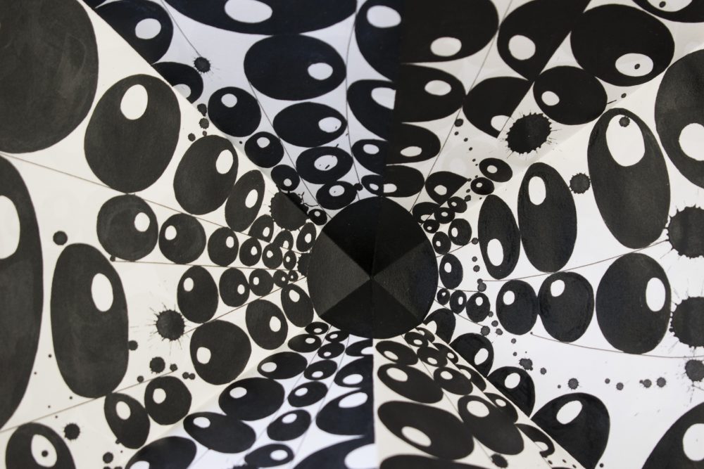Detail view of folded paper drawing, showing a black circle at the center and lines of black circles with small white circles inside them radiate outward from the center.