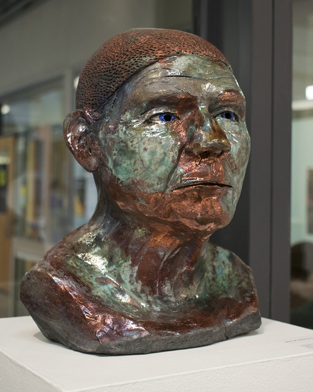 Ceramic sculpture of a bust of a head and shoulders, colored copper and green, sitting on a pedestal in front of an interior glass wall.