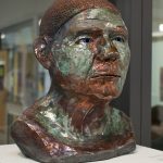 Ceramic sculpture of a bust of a head and shoulders, colored copper and green, sitting on a pedestal in front of an interior glass wall.