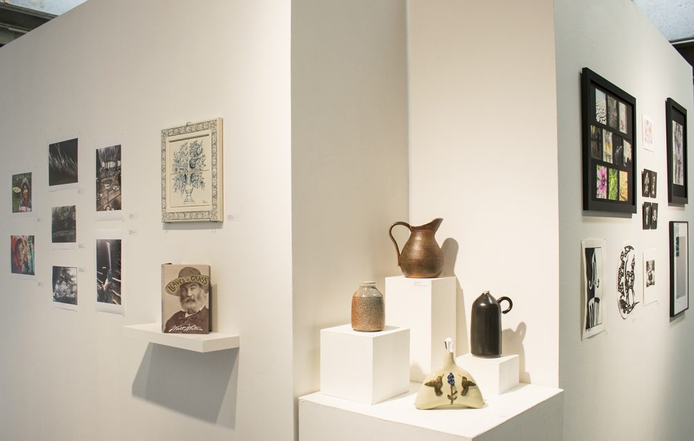 Installation view of gallery with a pedestal at the center and two gallery walls on either side. The pedestal has four ceramic sculptures; on the left wall are photographs, a book and a tile art piece; on the right wall are black and white drawings and three framed artworks.