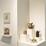 Installation view of gallery with a pedestal at the center and two gallery walls on either side. The pedestal has four ceramic sculptures; on the left wall are photographs, a book and a tile art piece; on the right wall are black and white drawings and three framed artworks.