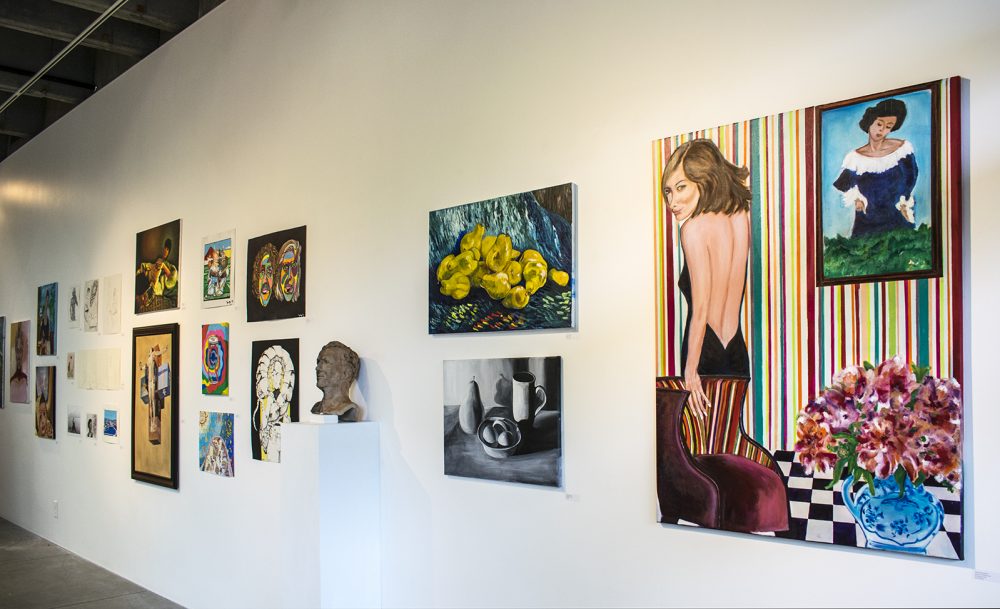 Installation view of gallery wall with multicolored and black and white paintings and drawings on a wall that recedes to the left. At the center of the wall is a ceramic sculpture of a male head.