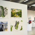 Installation view of a gallery with four multicolored paintings on paper on a wall in the foreground on the left. To the right, in the background are many abstract and representational paintings on the wall and several small ceramic sculptures on pedestals.