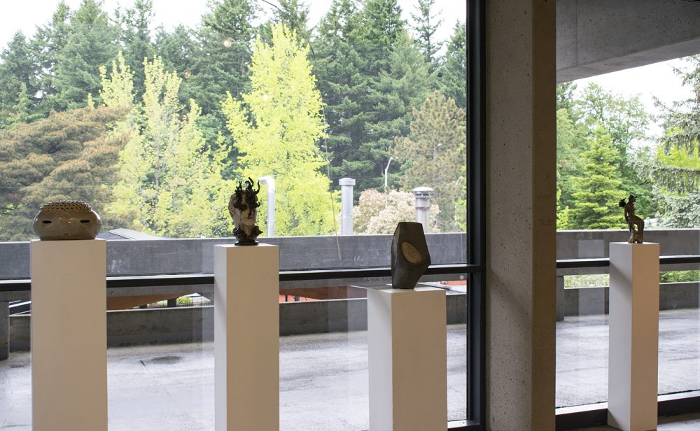 Installation view of a gallery with four pedestals in a row with small ceramic sculptures placed atop each one. Behind the pedestals are floor to ceiling windows that show an outdoor walkway and green trees beyond.