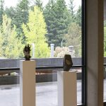 Installation view of a gallery with four pedestals in a row with small ceramic sculptures placed atop each one. Behind the pedestals are floor to ceiling windows that show an outdoor walkway and green trees beyond.