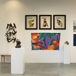 Panorama installation view of art gallery with many artworks on the far wall in the background, on additional walls to the left and rights, and of several pedestals in the middle.