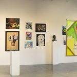 Panorama installation view of gallery with several artworks on the far wall in the background, on walls on the left and right, and on pedestals throughout the room. To the left is a freestanding artwork with windows behind it.